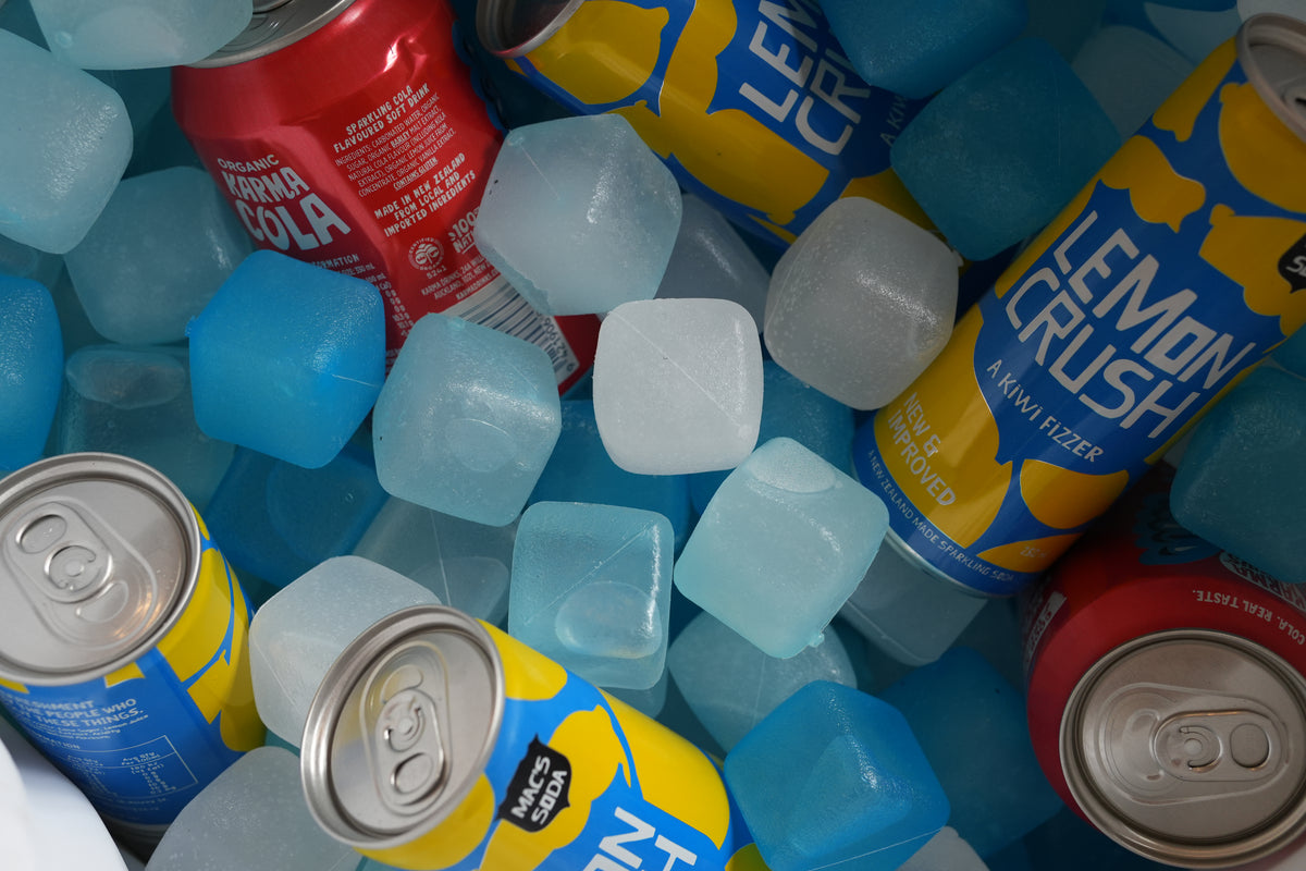 A close-up arrangement of canned sodas and lemonade frozen in blue ice cubes. Visible brands include Lemon Crush, Coca-Cola, and Fanta.